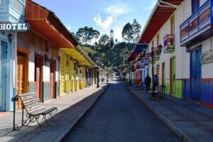 A narrow street lined with colorful buildings and a hotel sign on the left side. Brightly painted facades in yellow, red, and blue adorn the property in Colombia. Benches are placed along the sidewalk, and a hill with trees is visible in the background under a partially cloudy sky.