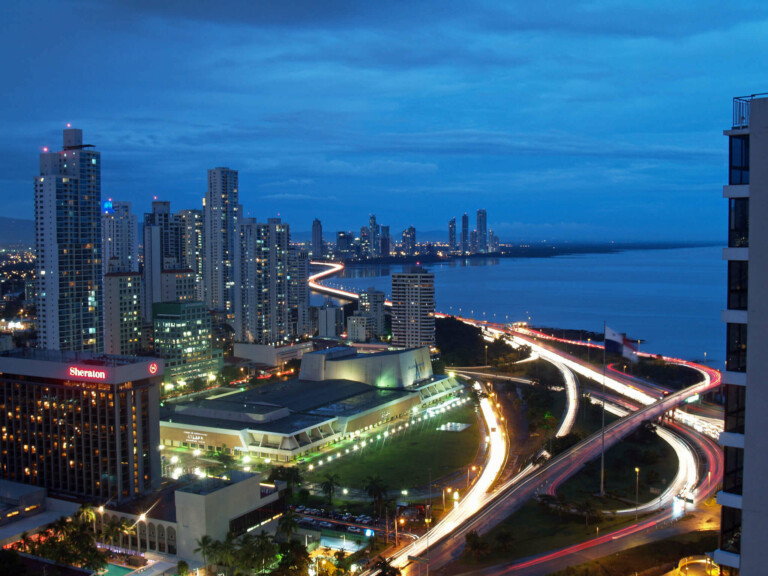 Un paisaje urbano nocturno que presenta un horizonte de edificios altos con luces encendidas. Una carretera sinuosa con rayos de luz de vehículos en movimiento discurre a lo largo del paseo marítimo. En primer plano se ven un hotel Sheraton que celebra su aniversario y un gran edificio moderno. El cielo está mayormente despejado.