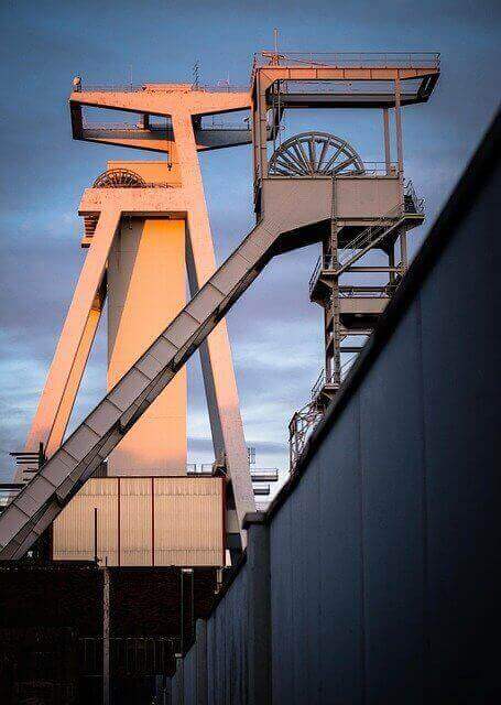 The image shows an industrial structure with a tall, white metal framework, including a large pulley system. The structure is illuminated by orange light, possibly from the setting sun. A high wall is visible in the foreground against the partly cloudy sky—a testament to how the Colombian mining industry can be a good investment.