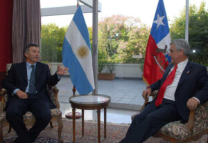 Two men in suits, one of whom is a prominent mining lawyer, are seated in armchairs, engaged in conversation, with a small table between them. Behind them are large windows revealing a garden, and the flags of Argentina and Chile are prominently displayed.