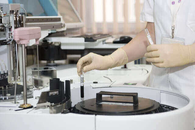 A technician wearing a white lab coat and latex gloves is handling a test tube inside a laboratory. Among the various pieces of lab equipment on the counter, including a centrifuge and other analytical devices, you can spot items used for testing medical marijuana.