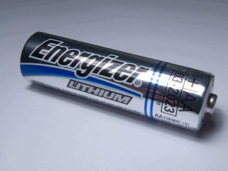 Close-up of a single Energizer branded lithium AA battery lying on a flat surface, resembling a tool from a mining lawyer's desk. The battery has a metallic silver and black casing with blue accents, displaying the "+" and "-" signs. The expiry date marked on it is "03.2023".