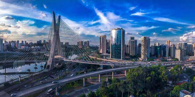 A panoramic view of a modern city with a prominent cable-stayed bridge over a river. The skyline features numerous high-rise buildings, including glass-covered towers, hinting at the mining wealth powering growth. The scene is set under a bright blue sky with scattered clouds and greenery in the foreground.