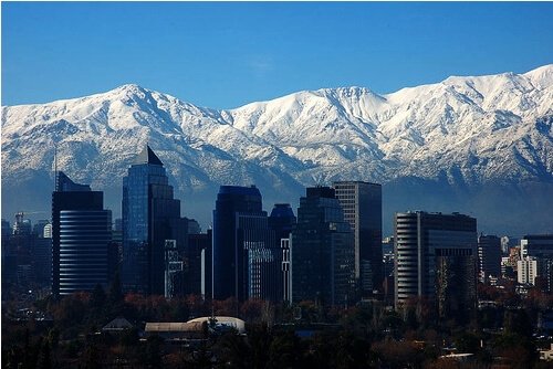 A cityscape with modern high-rise buildings stands in the foreground, while the snow-capped Andes mountains rise prominently in the background under a clear blue sky, showcasing Chile's booming startup sector.