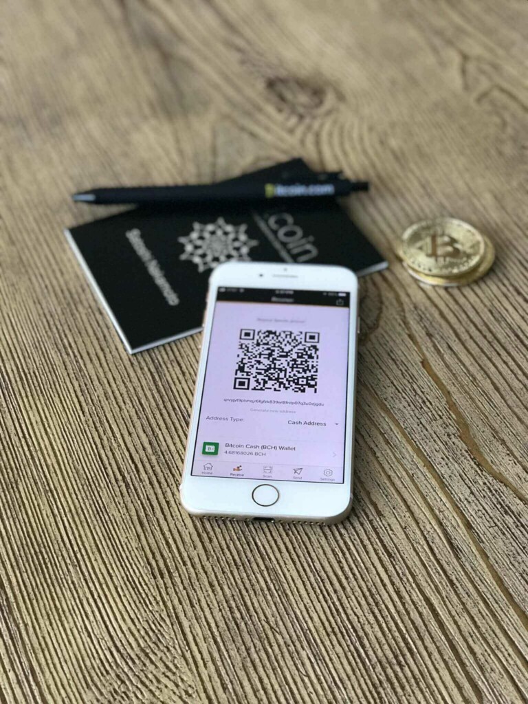 A smartphone displaying a QR code is placed on a wooden surface. Behind the phone, there is a black pen with white text and a small black booklet. To the right, a physical representation of a Bitcoin coin, symbolizing fintech innovation, is visible.