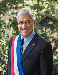 A person with gray hair is smiling, wearing a dark suit, blue tie, and a tri-color sash in red, white, and blue. Trees and foliage form the background. The attire suggests a role in Chile's governmental immigration reform efforts.