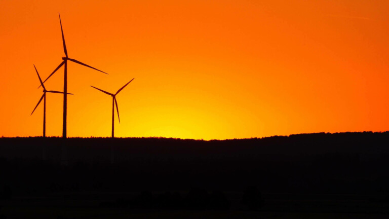 Two wind turbines stand against a vivid orange sky during sunset in Chile, with the sun partially visible on the horizon behind a dark, silhouetted landscape. The turbines are stationary, highlighting the contrast between the bright sky and dark foreground—a picturesque reminder of renewable energy's business opportunities.