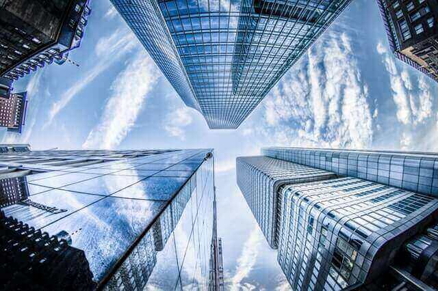 A fisheye perspective image captures several tall, modern skyscrapers with glass facades extending into the sky in Colombia. The buildings, potentially housing corporations like SAS, reflect the blue sky filled with scattered clouds, creating a dramatic and symmetrical urban scene.