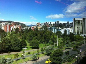 A cityscape with tall buildings under a blue sky, featuring a large green park with trees in the foreground. A yellow car and a few other vehicles are visible on the nearby road, capturing the vibrant hustle of Formação da Empresa.