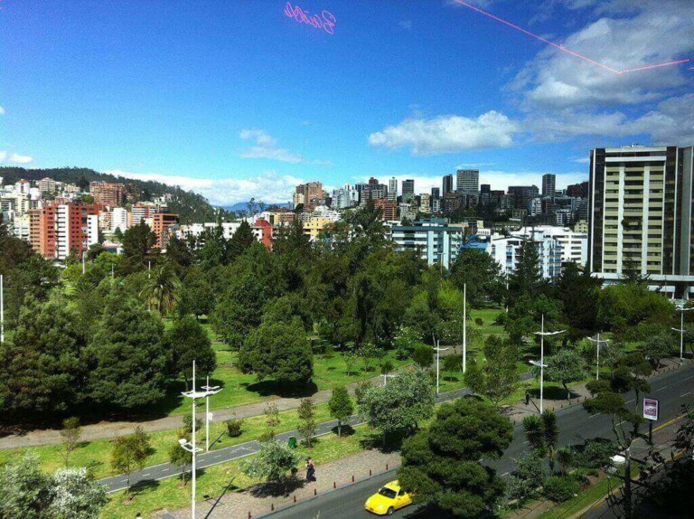 A cityscape with tall buildings under a blue sky, featuring a large green park with trees in the foreground. A yellow car and a few other vehicles are visible on the nearby road, capturing the vibrant hustle of Formação da Empresa.