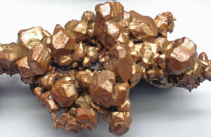 A cluster of shiny, reddish-brown crystallized copper mineral specimens is shown on a light grey background. The copper crystals have a metallic luster and are unevenly shaped, with some larger and some smaller formations intertwined. Such unique specimens spark interest not only among geologists but also in Mining Legal Due Diligence.