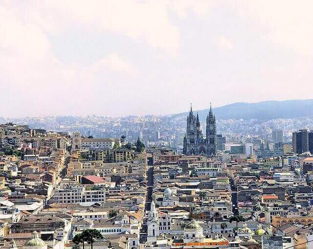 Aerial view of a city with clustered buildings and streets, featuring two prominent cathedral towers in the center. The background showcases rolling hills and a hazy sky, reminiscent of a scene you might find if looking for a Quito employment attorney amidst the cityscape.