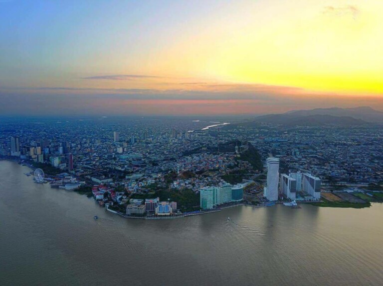 Aerial view of a coastal city at dusk, with tall buildings and an expansive urban area extending inland. The sun is setting behind the mountains in the background, casting a warm glow over the sky and water. Nestled along this coastline, business in Ecuador thrives as companies set up an export hub amidst greenery and developed areas.