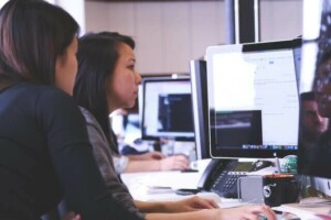 Dos personas están sentadas en un escritorio, mirando juntas la pantalla de una computadora en un ambiente de oficina. Parecen estar concentrados en el contenido que se muestra en el monitor, tal vez planeando establecer una compañía en Ecuador. Sobre el escritorio se ven varios objetos de oficina, como un teléfono, una taza y papeles.