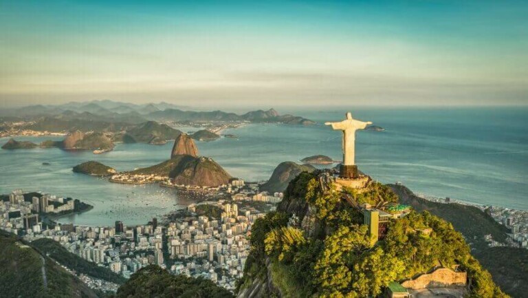 Panoramic view of the Christ the Redeemer statue atop a mountain, overlooking Rio de Janeiro. The cityscape includes densely clustered buildings near the coastline, hills, and the Atlantic Ocean with scattered islands in the background, offering a visual treat for those doing business in Brazil.