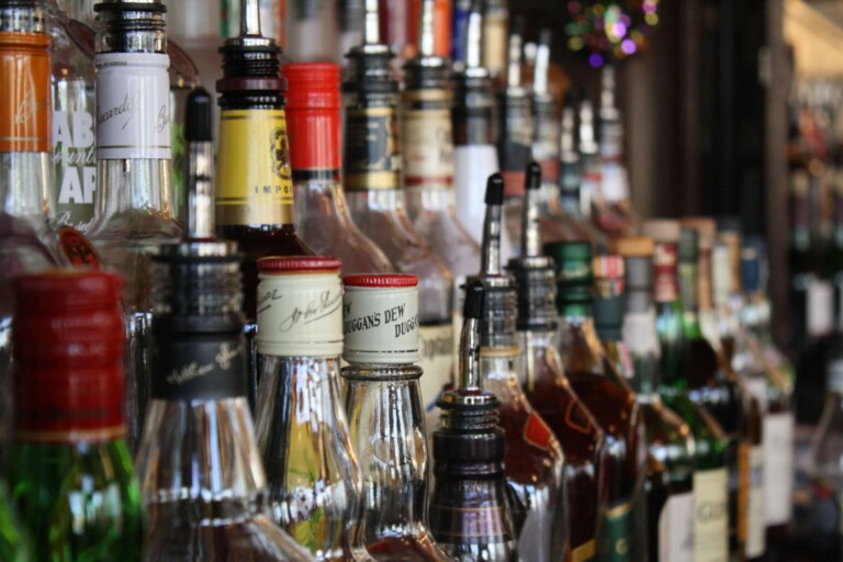 A close-up view of various liquor bottles lined up on a bar shelf. The bottles, representing the vibrant alcoholic beverage industry in Mexico, are of different shapes, sizes, and colors, and have labels indicating different brands of spirits. The background features more bottles and bar decor.