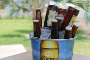 A bucket filled with ice and several bottles of Corona beer, a staple of the alcoholic beverage industry in Mexico, is placed on a table outdoors. The background features green grass and leafy plants, suggesting a casual outdoor setting.