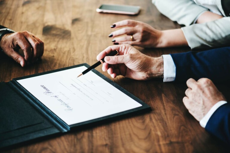 Tres personas están sentadas ante una mesa de madera. Una persona sostiene y apunta con un bolígrafo a un documento titulado "Certificado de matrimonio". Se ven las manos de otra persona con las uñas pintadas de negro. Al fondo, sobre la mesa, hay un teléfono inteligente que quizás contenga notas sobre la constitución de una empresa en Nueva Zelanda.