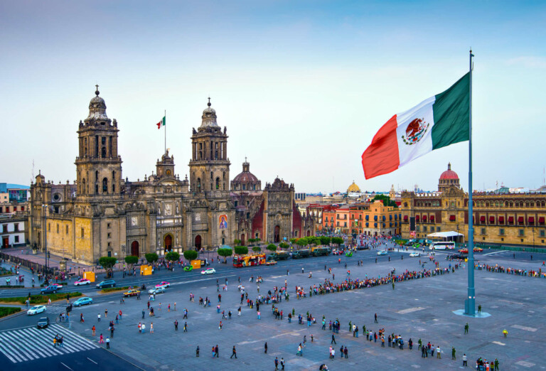 A large Mexican flag waves in the foreground of Zócalo Square in Mexico City. The Plaza, surrounded by historic buildings like the Metropolitan Cathedral, bustles with people and vehicles on nearby streets. The square's vibrancy makes it an iconic location for those doing business in Mexico.