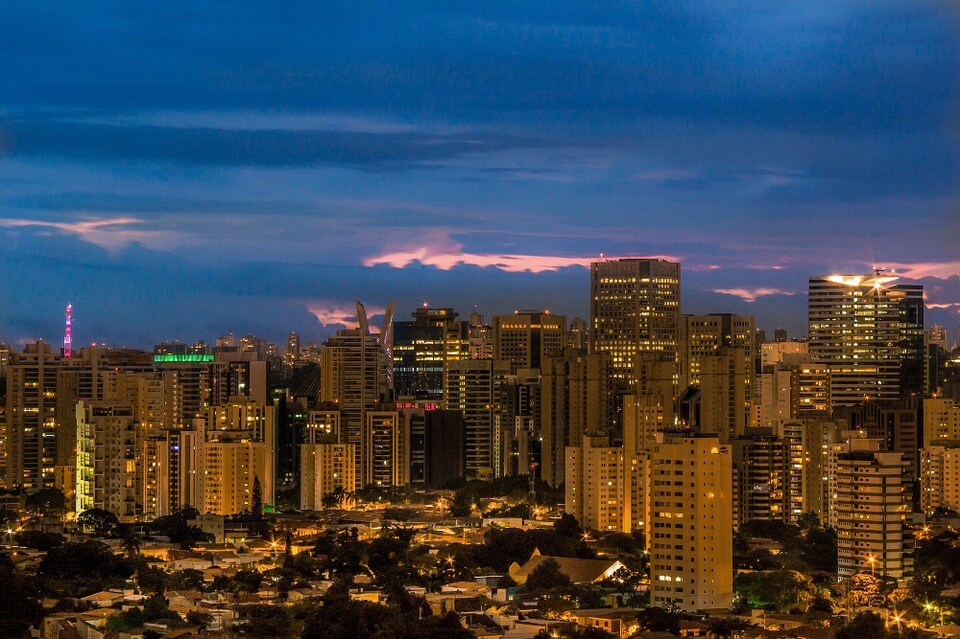 A cityscape at dusk featuring numerous tall buildings with lights starting to illuminate the scene, reminiscent of a bustling hub perfect for the formação da empresa. The sky displays a mix of dark blue and faint pink hues, while the foreground shows a blend of urban structures and patches of trees.