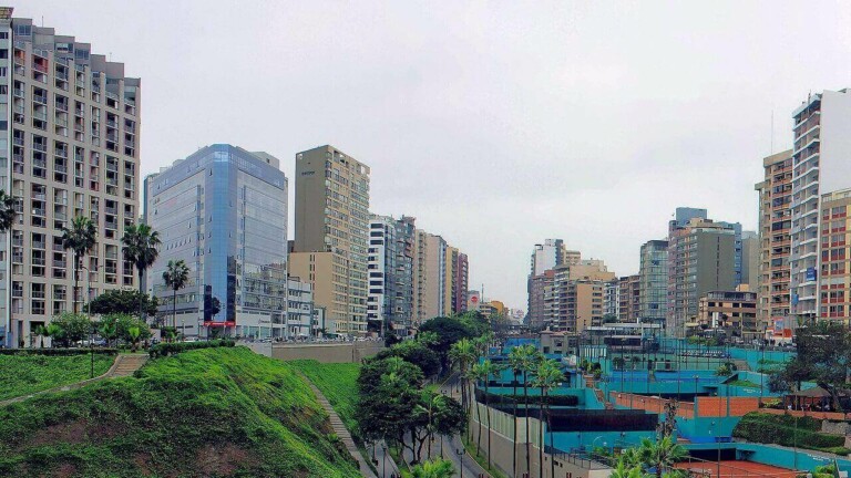 A view of a cityscape featuring tall residential and commercial buildings. Green grassy areas are visible on the left, while tennis courts and other recreational spaces are on the right. Amidst this bustling urban environment, plans to set up a branch in Peru are underway. The sky appears overcast.