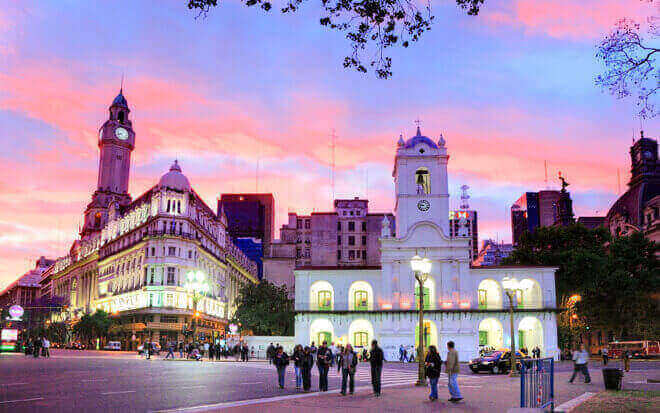 A bustling city square at twilight with a group of people gathering in front of an illuminated historic building, reminiscent of the diverse types of legal structures in Argentina. The sky is vivid with shades of pink, orange, and purple. Surrounding the square are various buildings, one featuring a prominent clock tower.