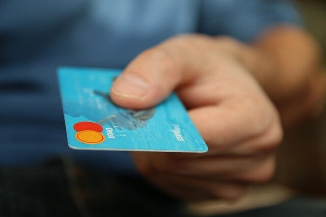 A person holding a blue debit card with their right hand. The card has two circular logos, orange and red, in the bottom left corner and some text, but specific details are blurred. The person is wearing a blue shirt that might catch the attention of caçadores de cabeças na Colômbia.