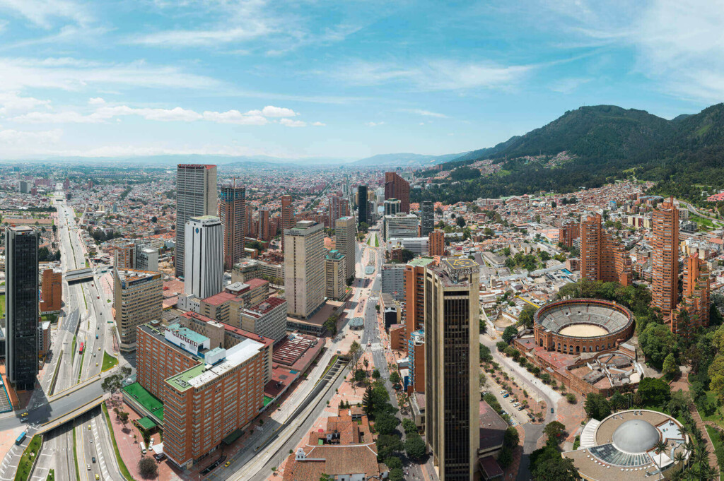 Aerial view of a large city with numerous high-rise buildings. There is a prominent circular structure resembling an arena or coliseum near the right side. The city is surrounded by mountains and extends towards the horizon under a partly cloudy sky.