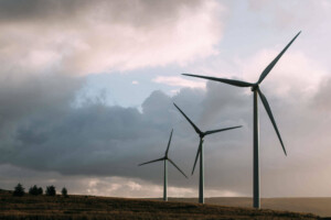 Three wind turbines are spread out along a grassy hill under a cloudy sky. The turbines, harnessing energy efficiently, stand tall with their blades angled in different directions. The landscape is mostly open, with a few small trees visible in the distance.