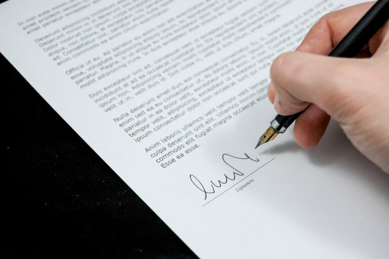 A hand holding a black fountain pen is signing a document at the bottom. The document contains several paragraphs of text in a formal layout, and the section being signed is labeled "Signature." It's reminiscent of agreements made by caçadores de cabeças na Colômbia, marked with precision and formality.