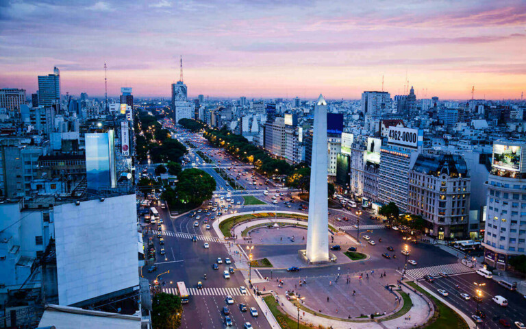 Vista aérea de una ciudad al atardecer, con un gran obelisco blanco en el centro rodeado de carreteras y edificios. Se ven automóviles en las carreteras y el horizonte incluye varios edificios de gran altura bajo un cielo rosa y azul, que recuerda a los bulliciosos centros metropolitanos de Argentina.