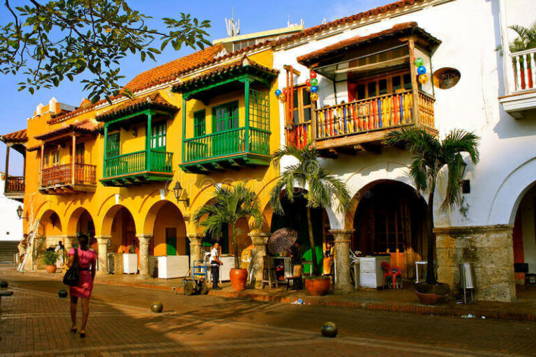 A street scene with colorful colonial-style buildings featuring arched doorways and vibrant balconies. A few people are visible, including a woman in a pink dress walking on the cobblestone street near an Independent Local Company. Some trees and outdoor seating areas in front of the buildings complete the lively atmosphere.