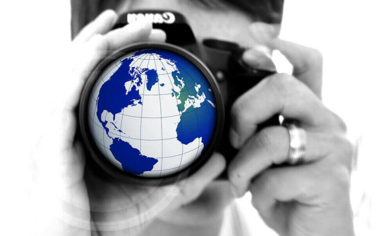 Close-up of a person holding a camera to their face, focusing on the lens. The camera lens displays an image of a globe with outlines of continents in blue, representing North America, South America, Europe, and parts of Africa. The image is in black and white except for the vibrant globe, symbolizing "Formação da Empresa.