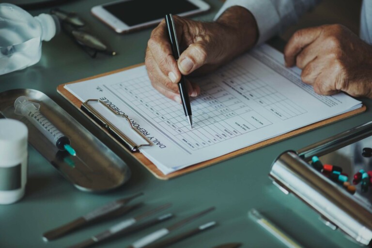 A person holding a pen fills out a daily report form on a clipboard. The table is scattered with various medical tools, including syringes, scissors, pills, and a smartphone. The person's attire suggests a professional setting related to the formação de empresa in the healthcare sector.