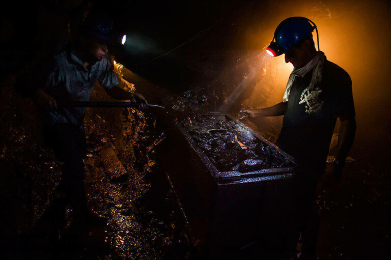 Dos mineros trabajan en una mina subterránea con poca luz. El minero de la izquierda está usando una herramienta para trabajar en algo dentro de un carro, mientras que el minero de la derecha, con un casco azul con un faro, está parado cerca del carro iluminado por una luz naranja. Esta escena capta la esencia de la inversión minera en Colombia y América Latina.