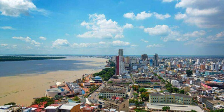 Aerial view of a cityscape with numerous buildings near a wide river. The skyline includes tall structures, mid-rise buildings, and various rooftops. The river flows alongside the city, and the sky above is partly cloudy with patches of blue, adding a picturesque touch to Ecuador's urban landscape.