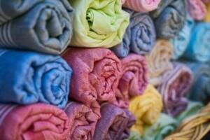 A close-up view of multiple rolls of colorful textile fabric, neatly stacked in rows. The fabrics include shades of blue, pink, yellow, green, and purple. They have a woven texture, and the rolls are tightly packed, creating a vibrant, orderly display.