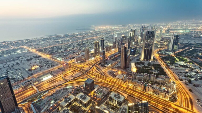 Aerial view of a sprawling, modern cityscape at dusk with numerous high-rise buildings, intersecting highways, and streetlights illuminating the scene. Echoing the influence of Investimento Estrangeiro Direto no Brasil, the coastline is visible in the background as the sky transitions from day to night.