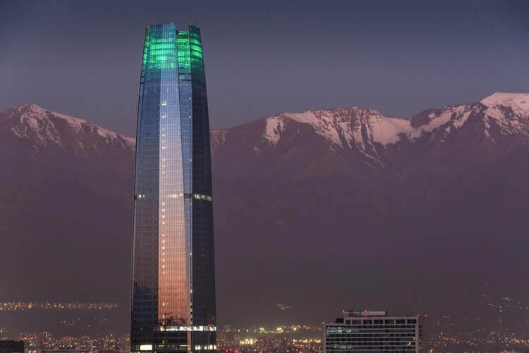 A tall skyscraper with a green-lit top stands against a backdrop of snow-capped mountains during twilight. Urban lights at the base of the building highlight the surrounding cityscape, a thriving hub for formação de empresa.