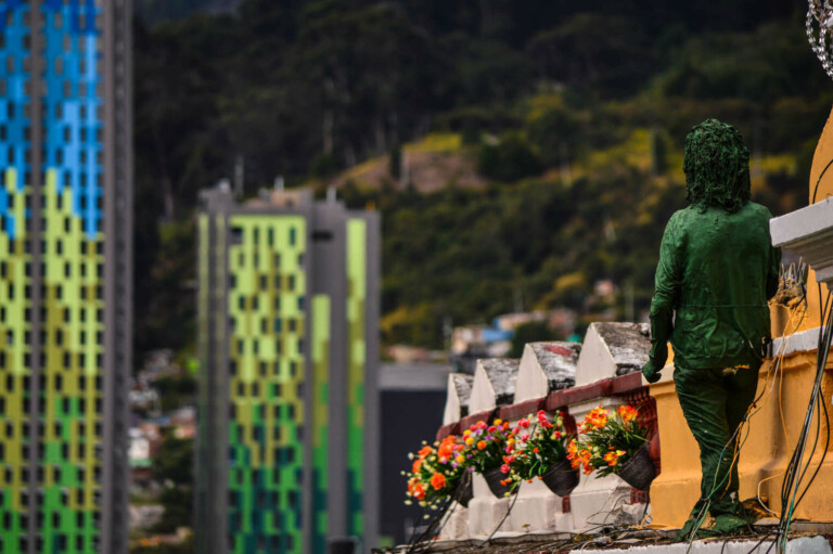 Una estatua verde se encuentra en una pared con vistas a los coloridos edificios del fondo. La pared está decorada con una hilera de macetas con flores y a lo lejos se ve una colina boscosa.