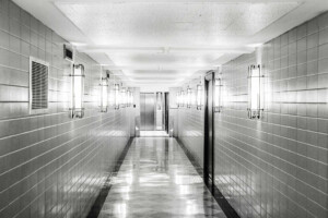 A black-and-white photo of a long, narrow hallway with tile walls and a reflective floor showcases the sleekness of ceramics. The hallway is lit by evenly spaced wall-mounted lights, leading to a closed elevator door at the far end.