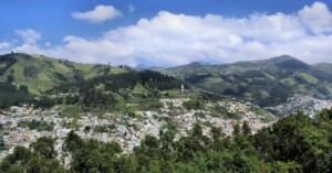 A landscape view of a city nestled among green hills under a partly cloudy sky. The city has numerous buildings spread across the valley and hillsides. The foreground features a dense forested area.