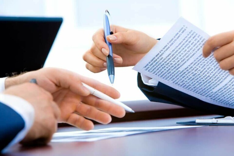 Two people in business attire hold pens while discussing and pointing at a document filled with text about Formação da Empresa. A laptop is partially visible in the background. One person's hand is extended, indicating emphasis on a particular section of the document.