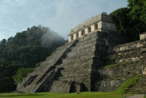 A large stone pyramid structure located at the base of a forested hill, partially shrouded in mist, evokes the rich history of ancient Mexico. The pyramid has steep stairs leading up to a temple or building at the top. The surrounding area is grassy, and the background is lush with dense foliage.