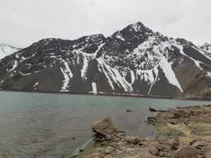 A mountain partially covered in snow stands by a lake with a rocky shore in the foreground under a cloudy sky. Snow patches are visible on the mountain slopes, and the water of the lake is a light blue-green color, offering an idyllic setting that promotes sustainable tourism near Santiago de Chile.