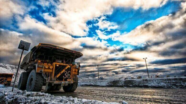 Un gran camión minero marcado con el número 36 está estacionado en un camino de tierra en un paisaje nevado. El cielo arriba está nublado con manchas azules. Al fondo se ven equipos industriales del sector minero en México.