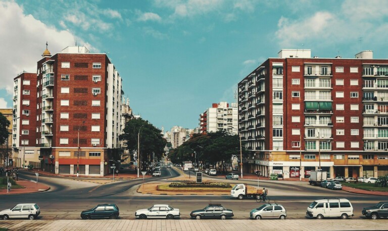 A city street with a median strip is lined with multi-story red brick buildings on both sides, giving it a classic look reminiscent of the early days of formação da empresa. Various vehicles, including cars and vans, are parked along the street and the median. The sky is partly cloudy, and there is light urban activity.