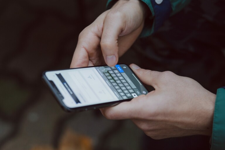 Una persona sostiene un teléfono inteligente con ambas manos y escribe en el teclado de la pantalla táctil. La pantalla del teléfono muestra una aplicación de mensajería, probablemente hablando de algún negocio o comercio electrónico. El individuo viste una chaqueta oscura con manga verde. El fondo es pavimento borroso.