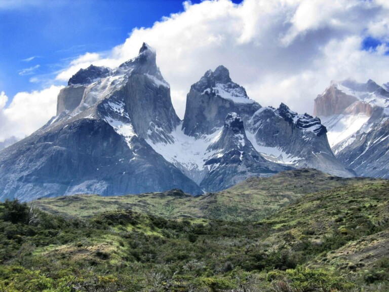 Los picos nevados bajo un cielo azul parcialmente nublado se elevan sobre un primer plano de vegetación verde. El terreno accidentado presenta crestas afiladas y dentadas y una mezcla de roca y vegetación.