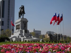 A statue of a man on a horse is mounted on a large stone pedestal in a city square. The area, vibrant with colorful flowers and several Chilean flags, stands as a reminder of the country's history. Tall buildings are visible in the background under a clear blue sky, symbolizing progress and modernity similar to VAT in Chile's development.